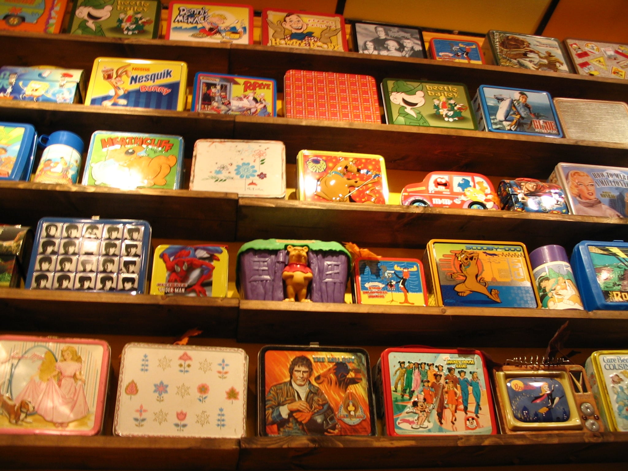 Colorful vintage lunchboxes with various cartoon and pop culture designs are displayed on wooden shelves, spanning across several rows in a bright, nostalgic collection.
