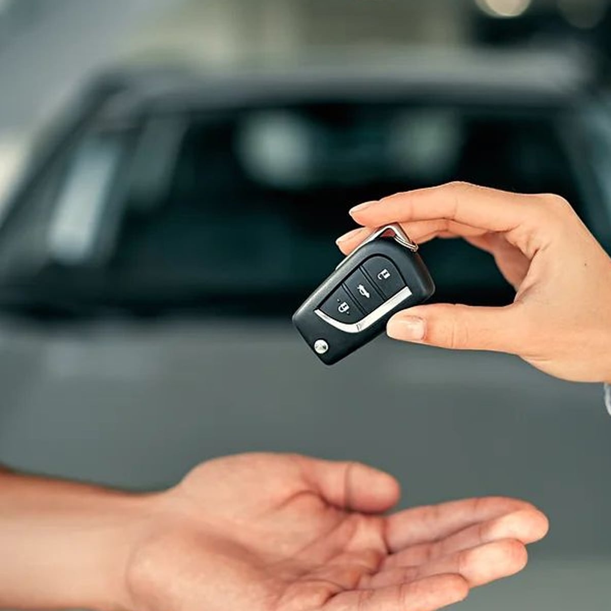 A close-up of one person handing a car key fob to another person, with a blurred car visible in the background. The receiving hand is open and facing up.