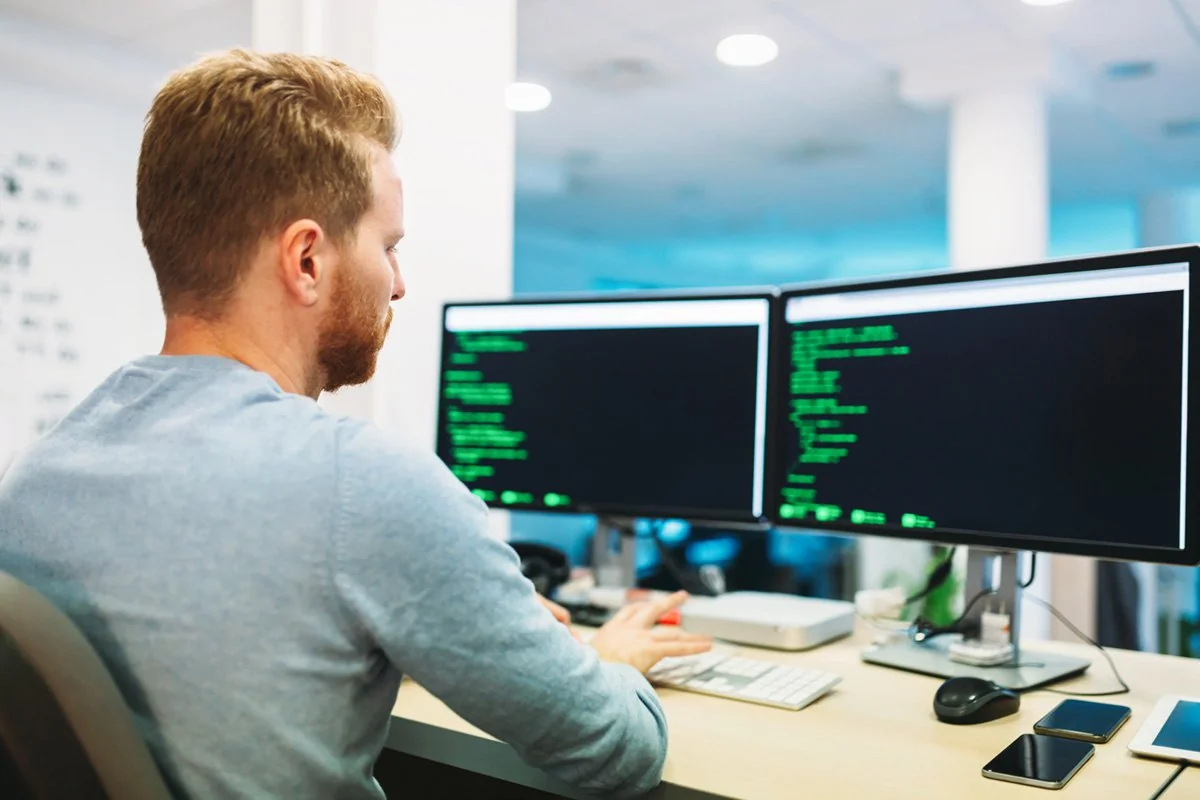 A person with short hair and a beard is sitting at a desk, working on two computer monitors displaying code or terminal screens. Several smartphones are placed on the desk beside the keyboard and mouse.