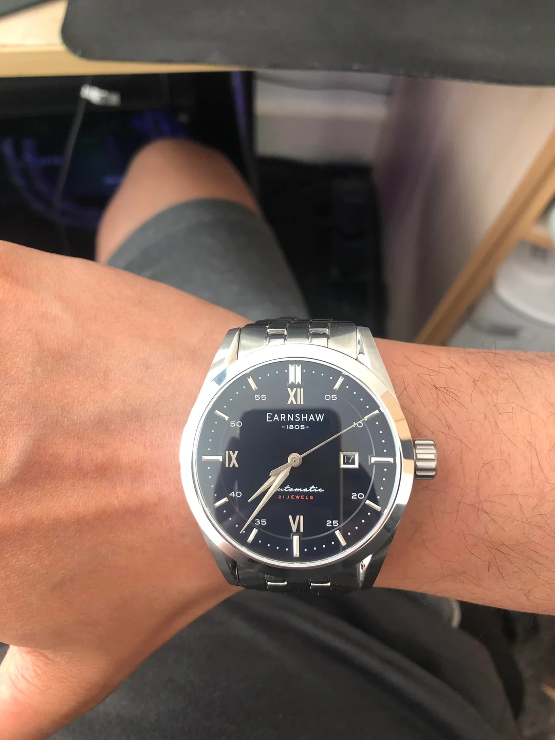 A person wearing a silver Earnshaw 1805 wristwatch with a black face, Roman numerals, and a metal band. The watch displays the date and time, and the person is dressed in gray shorts.