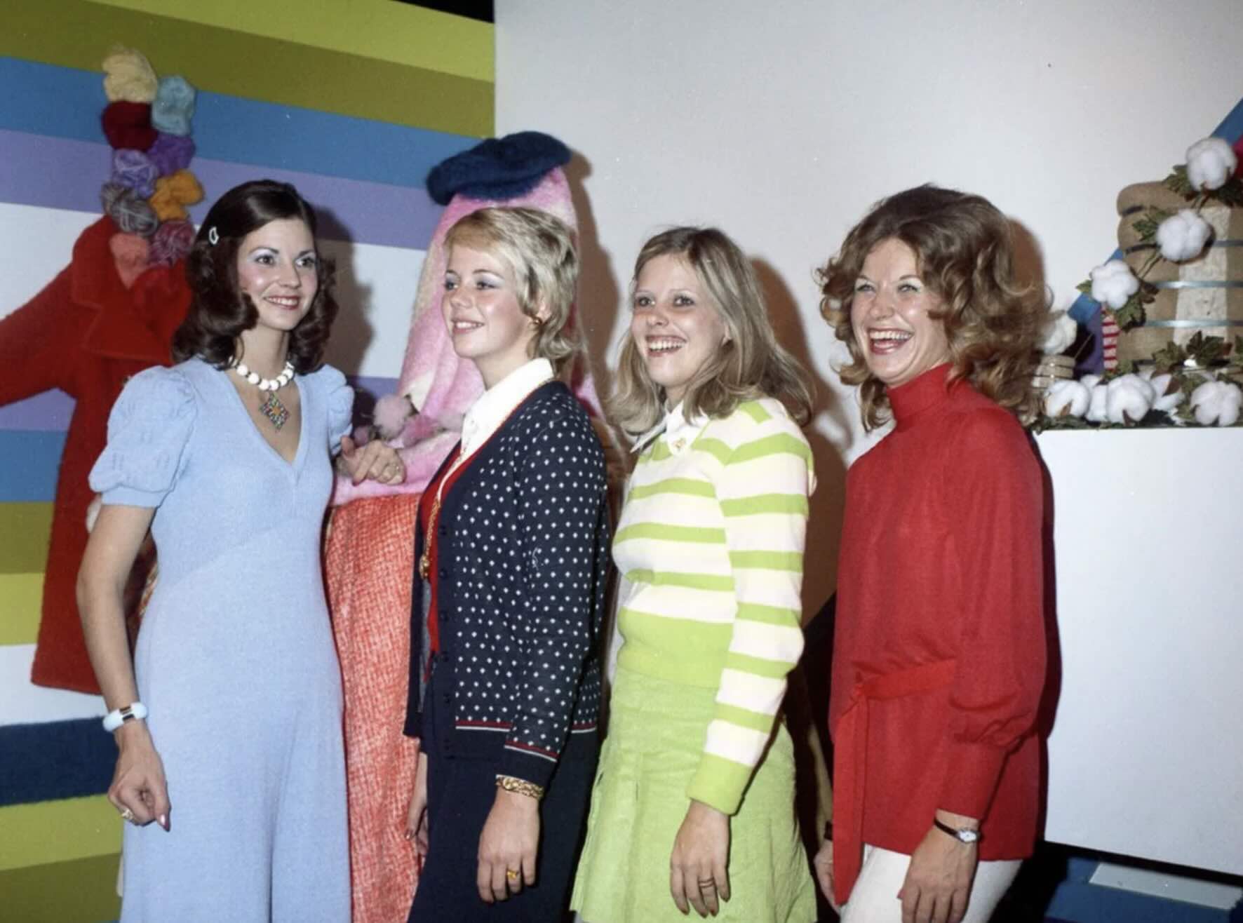 Four women stand smiling in front of a colorful, striped background with yarn and fabric decorations. They wear bright, fashionable 1970s-style outfits, including dresses, sweaters, and skirts.
