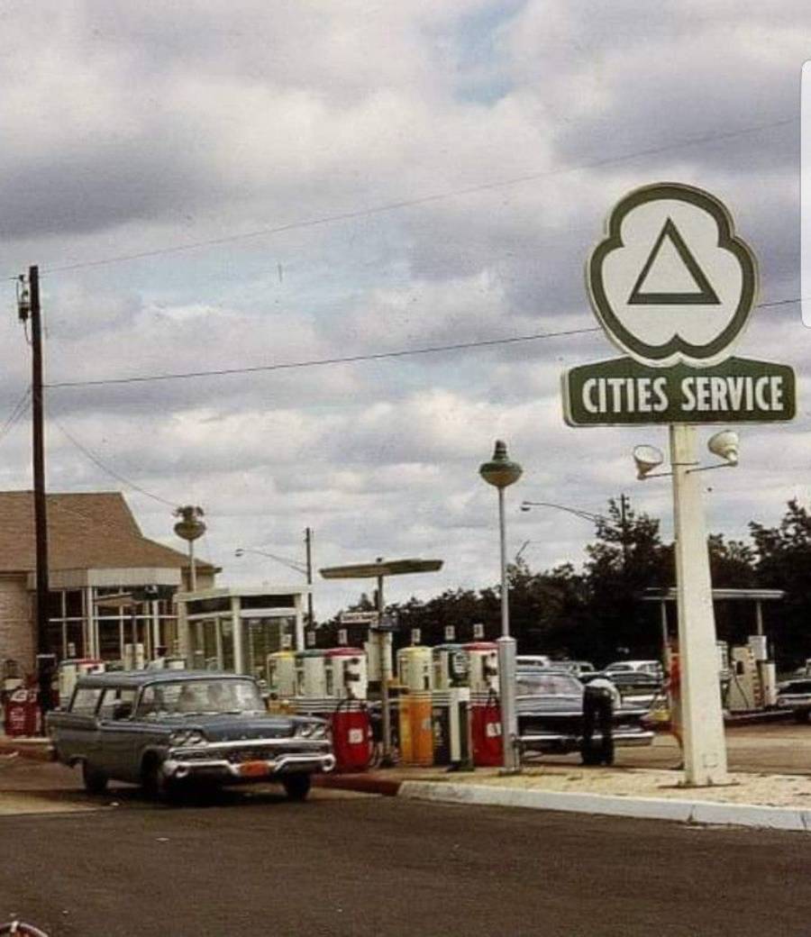 A vintage gas station with a "Cities Service" sign, several old-fashioned gas pumps, and classic cars parked nearby under a cloudy sky. The scene has a nostalgic, mid-20th-century atmosphere.