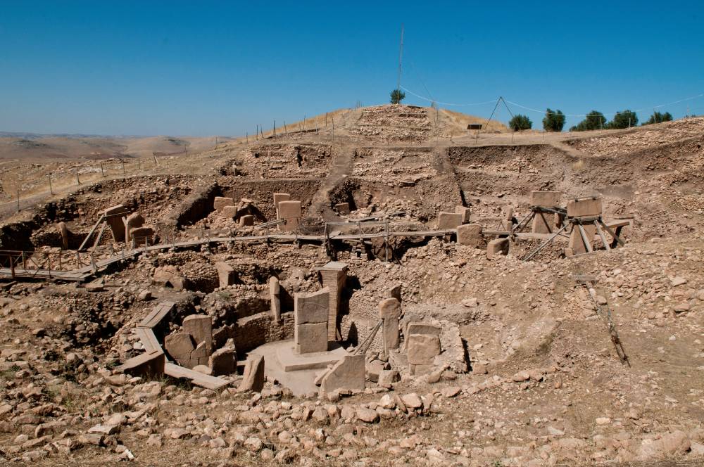 An archaeological site with large, ancient stone pillars and circular enclosures partially excavated in rocky, hilly terrain under a clear blue sky. Wooden walkways cross the site.