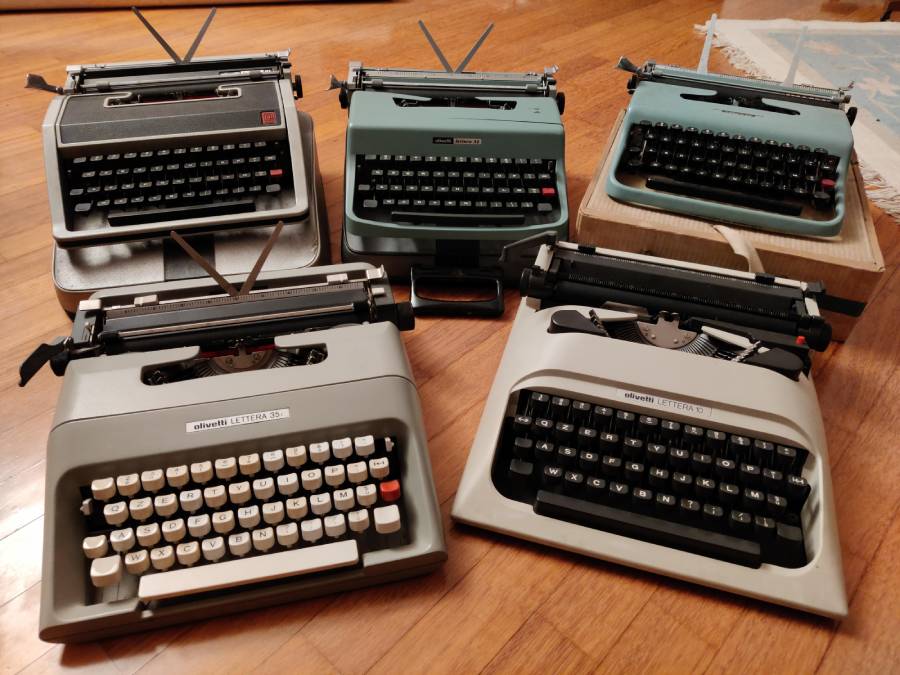 Five vintage typewriters of different colors and models are arranged on a wooden floor, with three in the back row and two in the front, showcasing their keys and design.