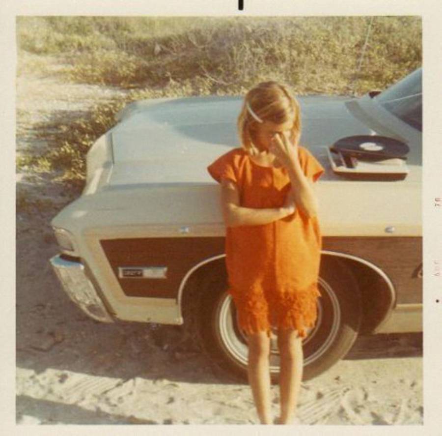 A young girl in an orange dress stands in front of a vintage car, covering part of her face with her hand. A vinyl record sits on the car’s hood. The scene appears to be outdoors on a sunny day.
