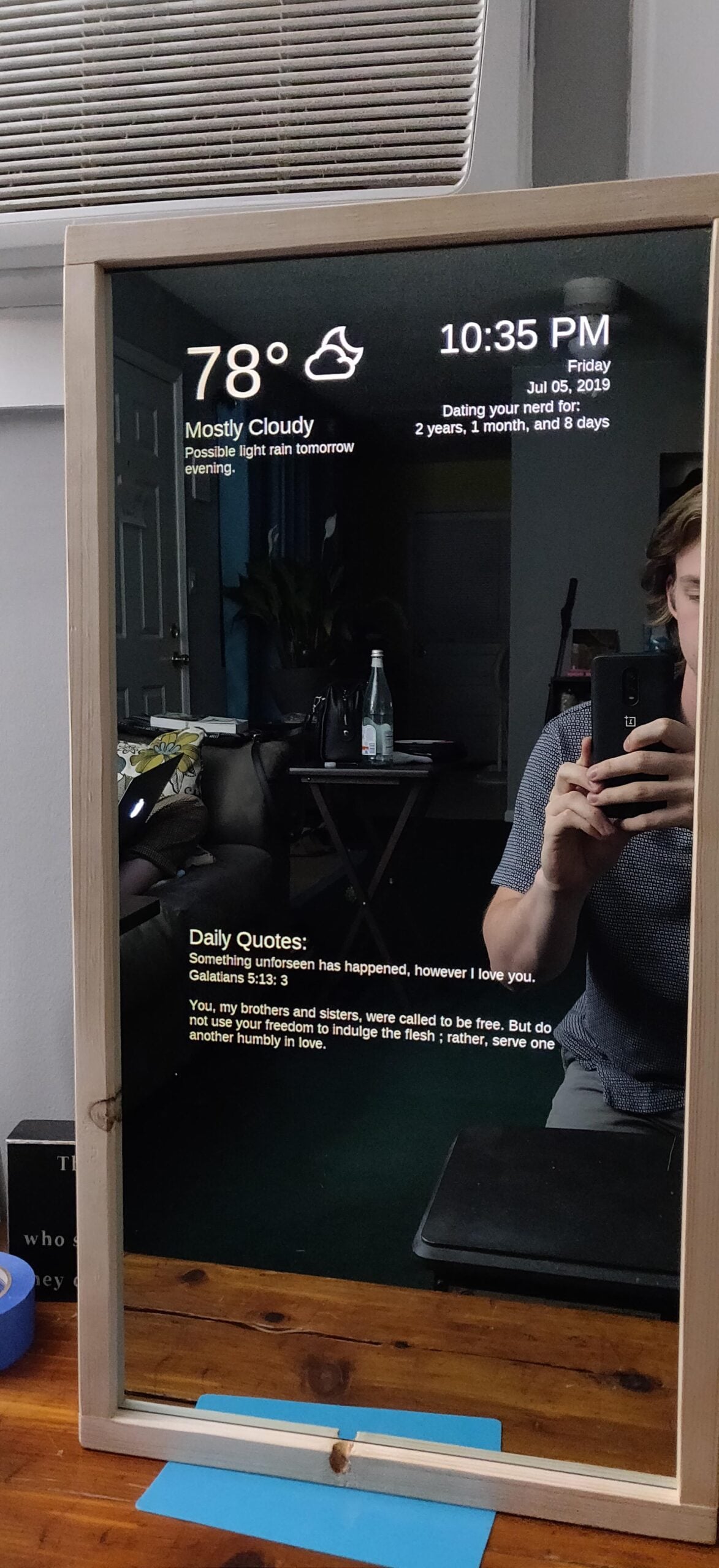 A smart mirror displays the weather (78°F, mostly cloudy), time (10:35 PM), date (Friday, July 5, 2019), and inspirational quotes. A person holding a phone is reflected, and a desk with various items is in the background.