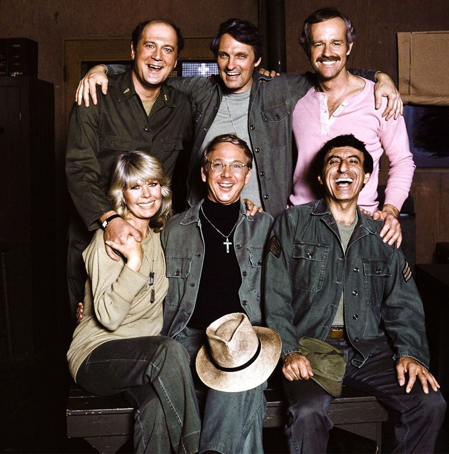 Six people from the TV show M*A*S*H sit and stand together indoors, smiling and wearing army-style clothing. One woman and five men pose closely, showing camaraderie and friendship.
