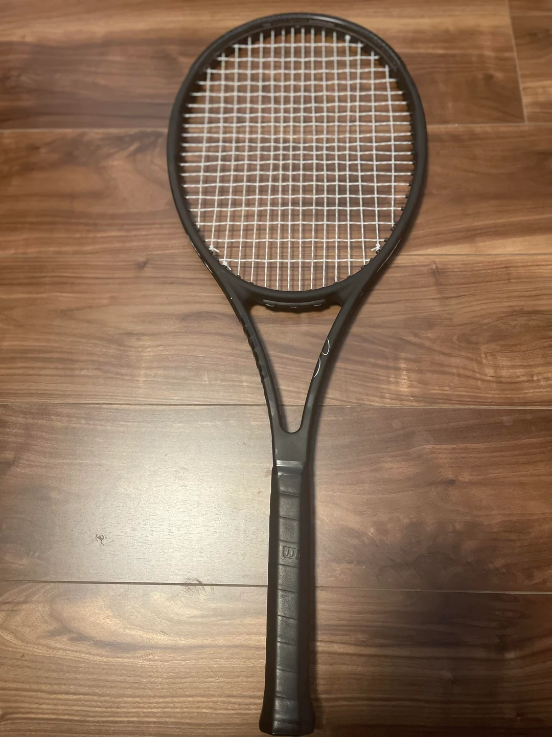 A black tennis racket with white strings is lying flat on a wooden floor.