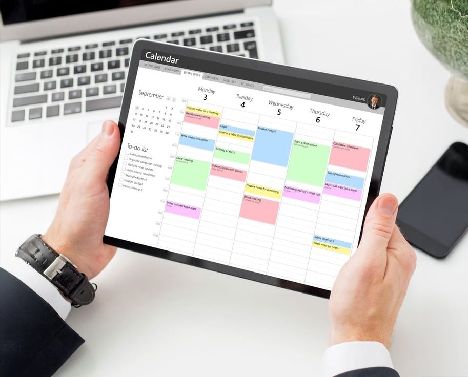 A person in business attire holds a tablet displaying a digital calendar for the week, with various color-coded events and a to-do list visible. A laptop and papers are on the desk in the background.
