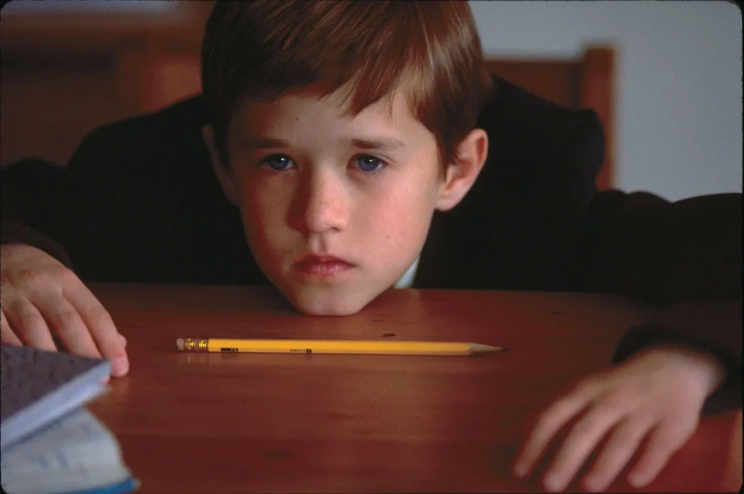 A young boy with brown hair sits at a wooden table, looking intently at a yellow pencil lying in front of him. His hands rest on the table, and an open notebook is visible to the side.