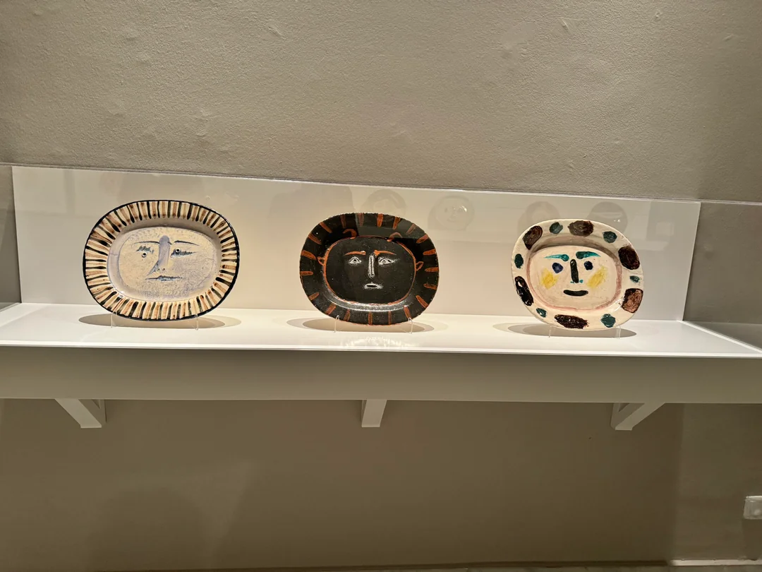 Three decorative plates with abstract, face-like designs in different colors and styles are displayed on a white shelf against a gray wall.