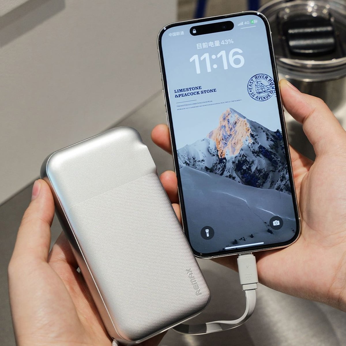 A person holds a smartphone displaying a mountain wallpaper and the time 11:16, while the phone is connected to a silver portable power bank with a charging cable.