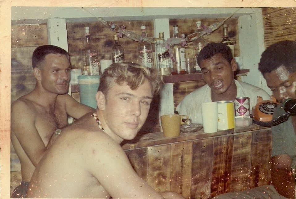 Four shirtless men sit closely together in a rustic bar with wooden walls, drinks, and liquor bottles behind them. They appear relaxed, with cups and a rotary phone on the table in front.
