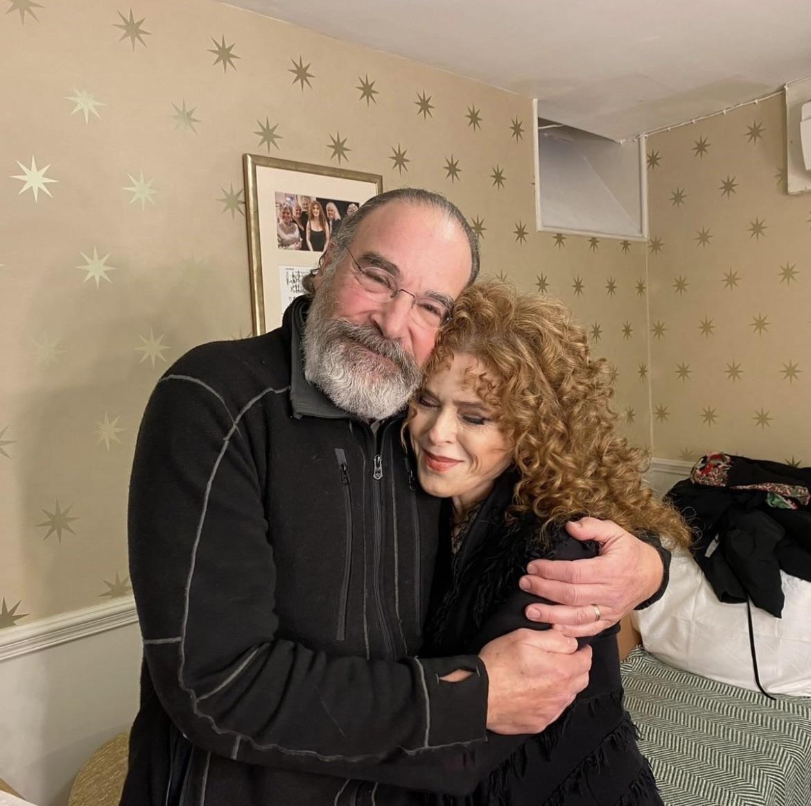 A bearded man and a woman with curly hair warmly hug each other and smile in a cozy, star-patterned room with framed photos and a bed in the background.