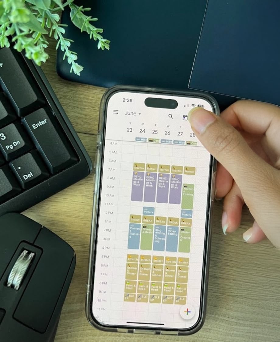 A hand holds a smartphone displaying a colorful, detailed weekly calendar. A keyboard, mouse, and a potted plant are on the wooden desk in the background.