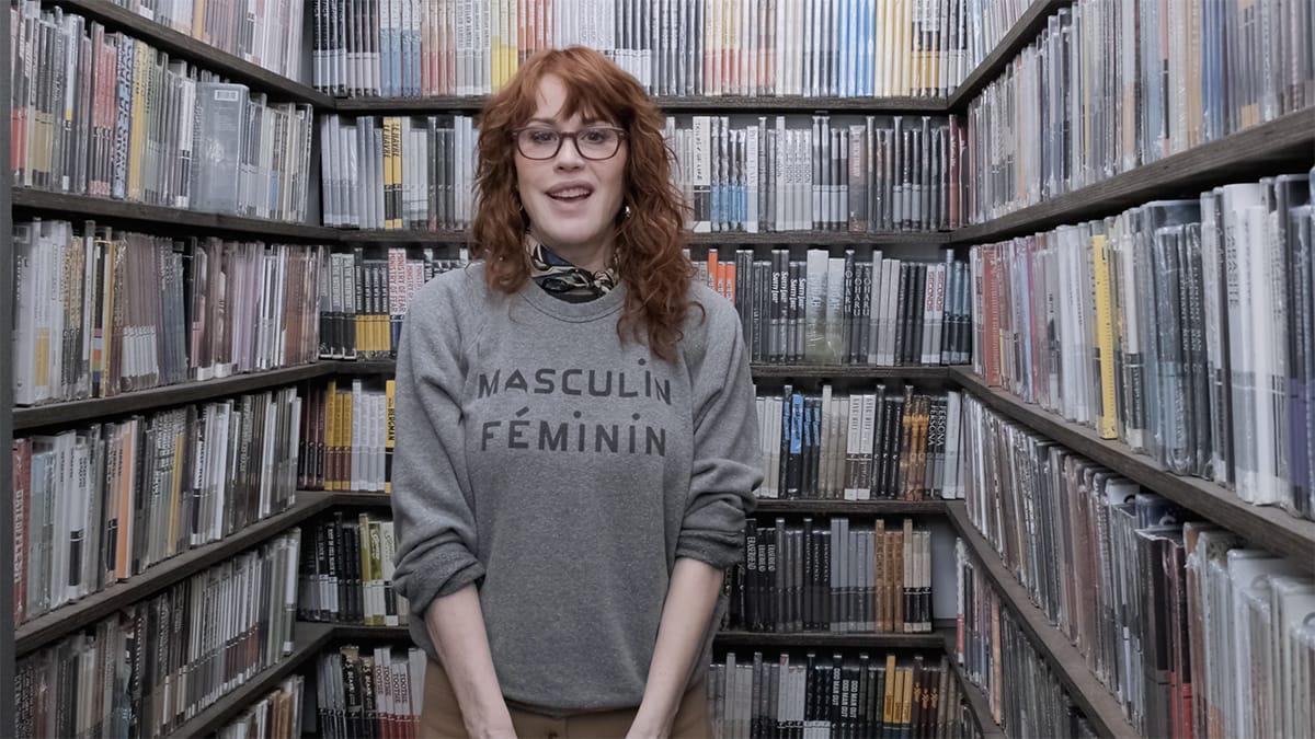 A person with curly red hair and glasses stands between shelves filled with books, wearing a grey sweater that says "MASCULIN FÉMININ.