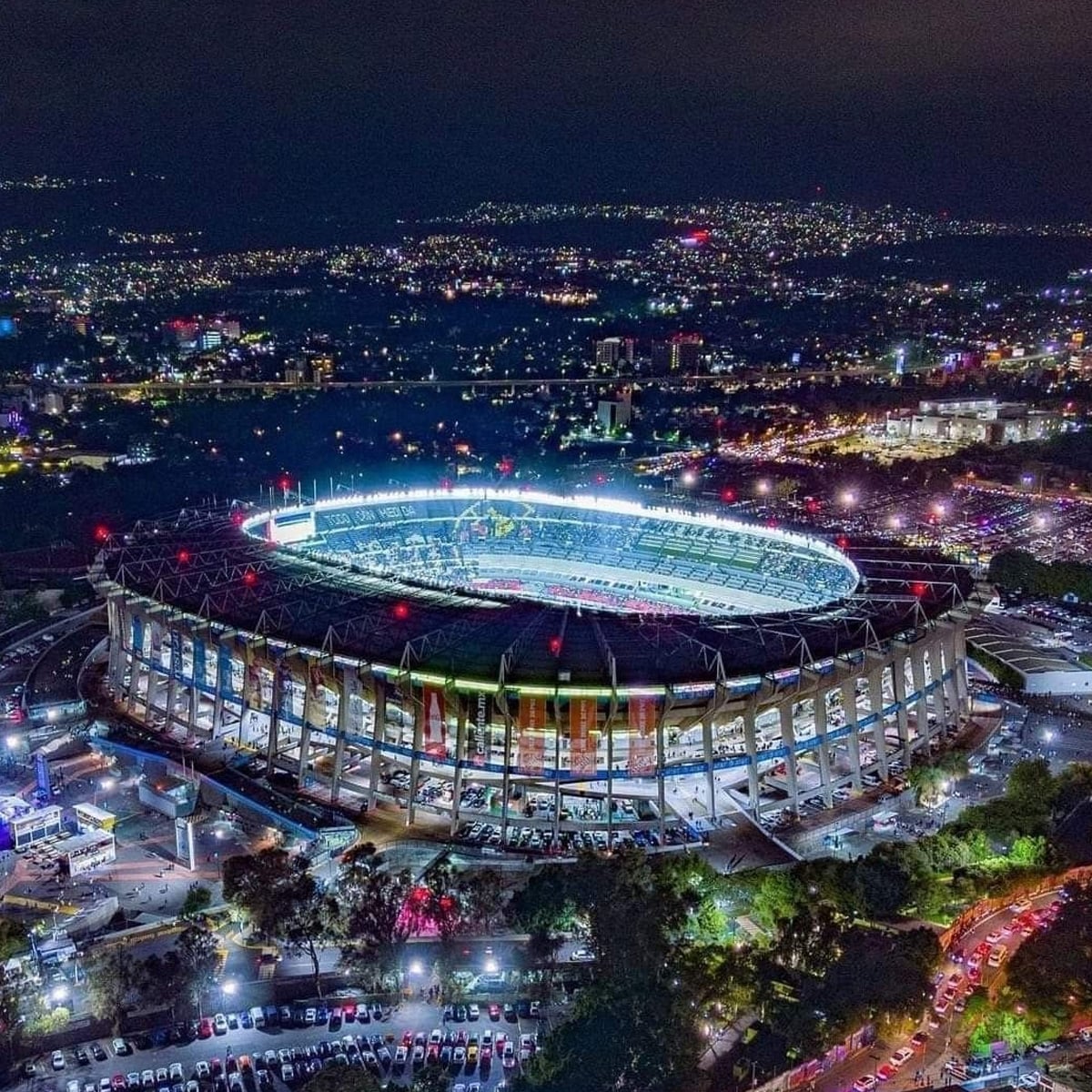 A brightly lit stadium at night, surrounded by trees and parking lots filled with cars, with a cityscape and twinkling lights in the background.