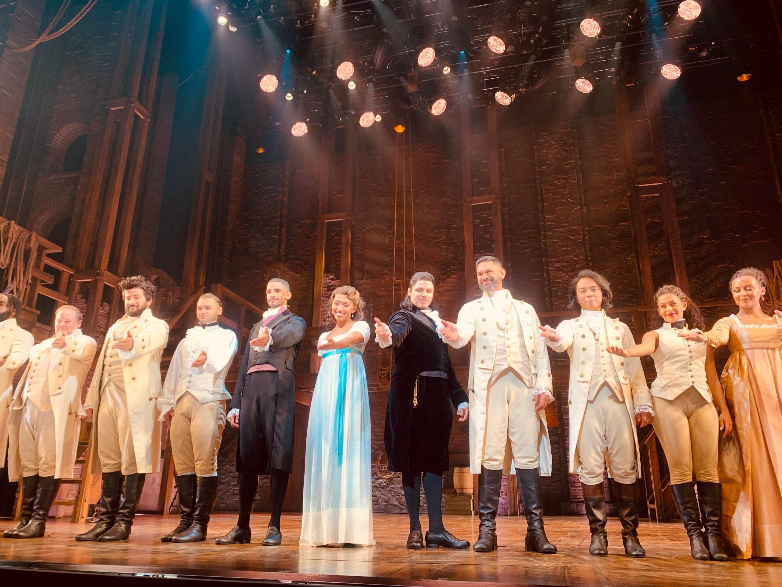 A group of actors in period costumes stand in a row on stage, holding hands and bowing to the audience under bright stage lights and a brick set backdrop.