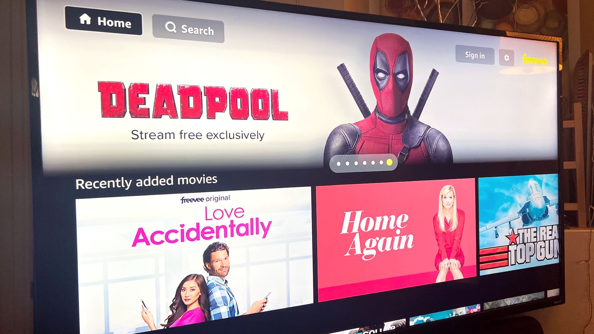 TV screen displays a streaming service menu featuring "Deadpool" as a highlighted title. Below, recently added movies include "Love Accidentally," "Home Again," and "The Real Top Gun.