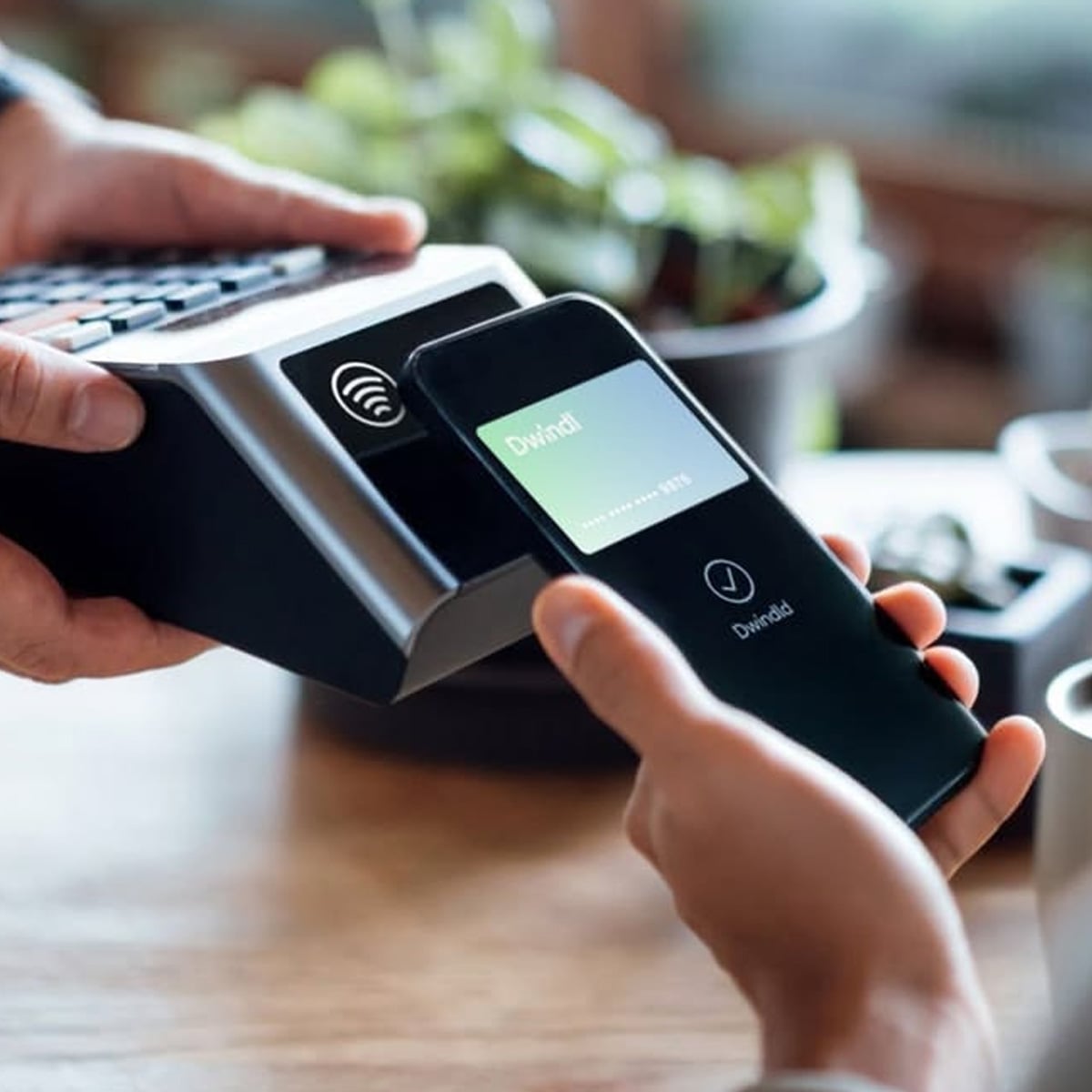 A person holds a smartphone close to a payment terminal, making a contactless payment. The phone screen displays a confirmation message with a check mark.