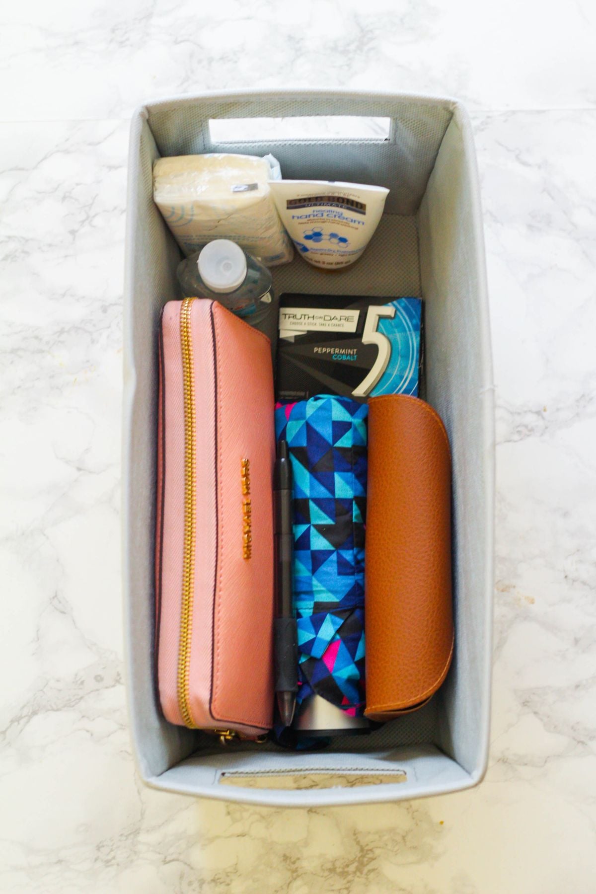 A gray storage bin containing a pink zippered pouch, a brown eyeglass case, a geometric-patterned pouch, a pack of 5 gum, a small water bottle, lotion, tissues, and a pen, all arranged neatly on a marble surface.
