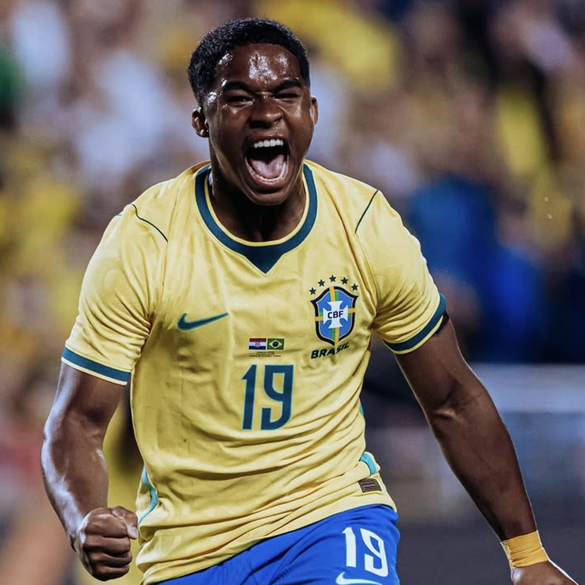 A Brazilian soccer player in a yellow jersey with number 19 celebrates passionately, clenching his fist and shouting, with a blurred crowd in the background.