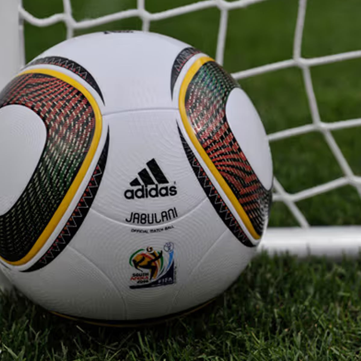 A close-up of an Adidas Jabulani soccer ball resting on green grass near a white goalpost, with a net in the background. The ball features colorful patterns and the 2010 FIFA World Cup South Africa logo.