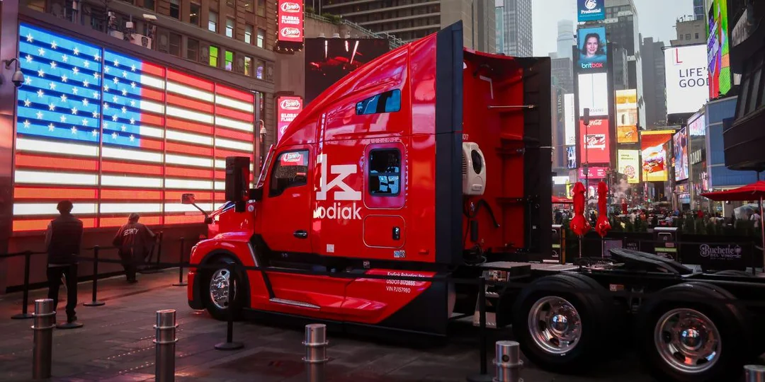 A bright red Kodiak semi-truck is parked in Times Square, New York City, near a large digital American flag display, with people walking nearby.