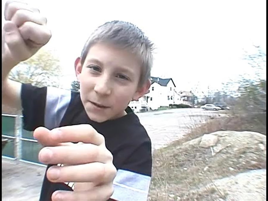 A young boy with short hair stands outdoors, raising his fists playfully toward the camera as if pretending to box. Houses, a parked car, and bare trees are visible in the blurred background.