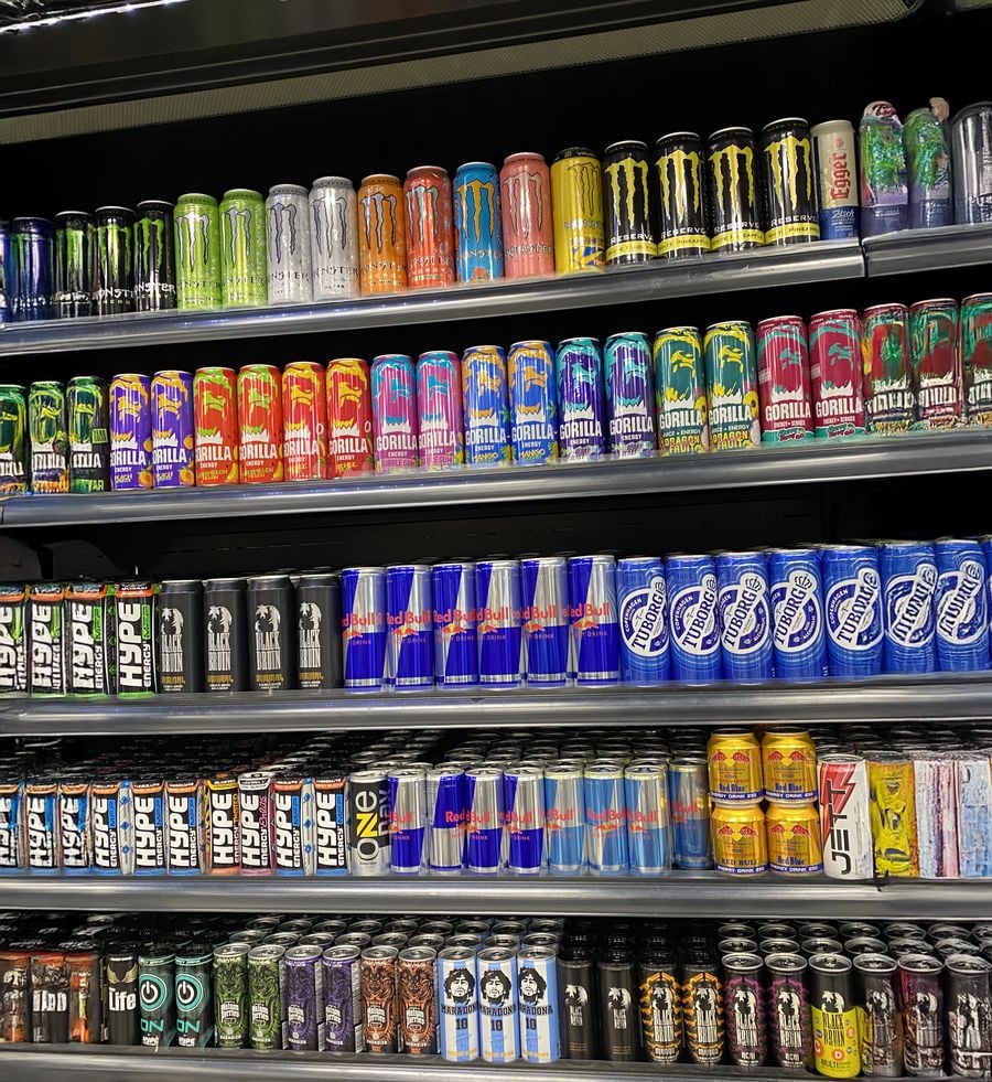 Shelves stocked with a colorful variety of energy drink cans, including brands like Monster, Red Bull, and others, organized by color and brand in a refrigerated display at a store.