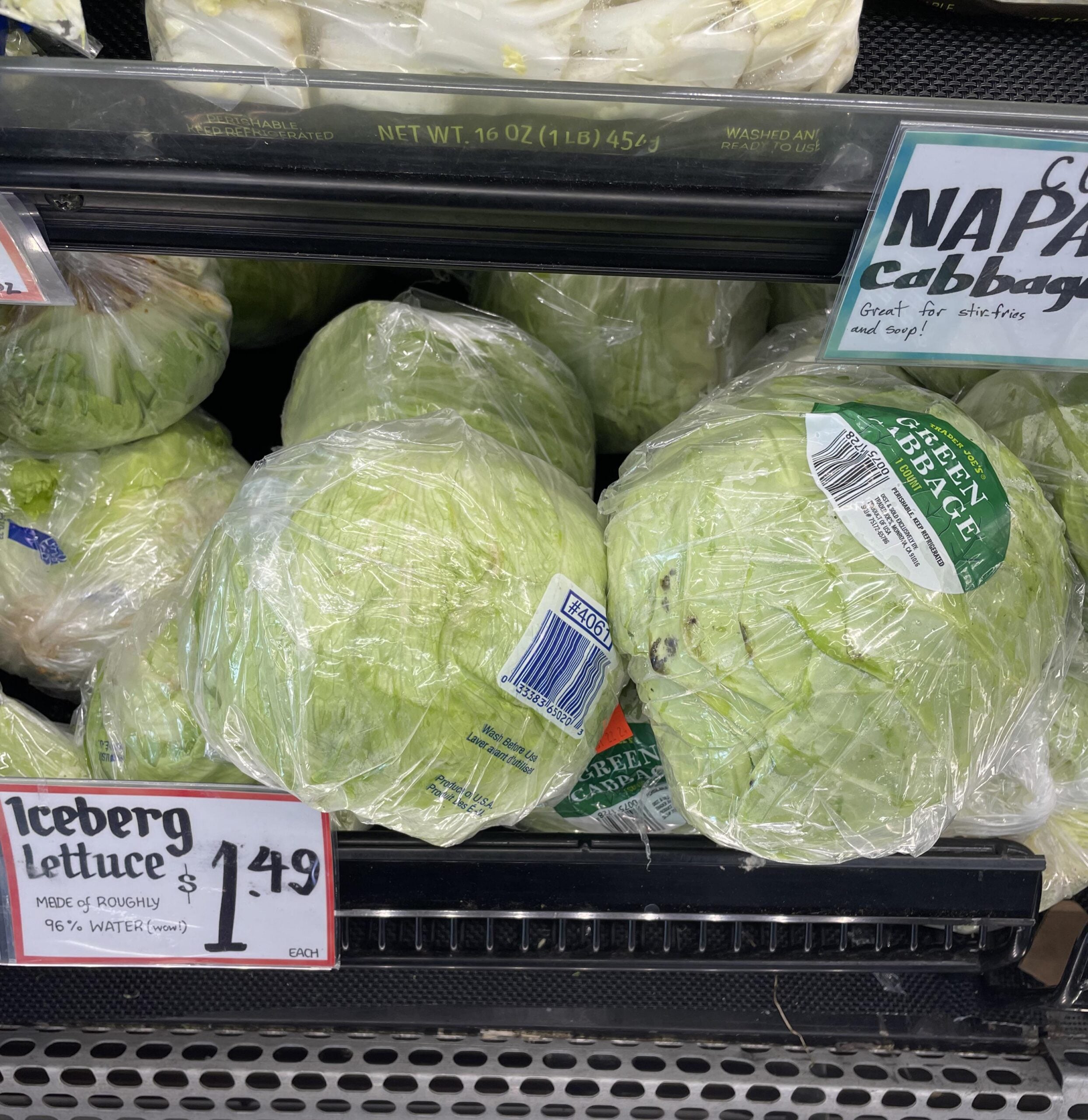 Several heads of iceberg lettuce wrapped in plastic are displayed on a grocery store shelf. A price sign in front reads "Iceberg lettuce $1.49." Nearby, a partial sign for Napa cabbage is visible.