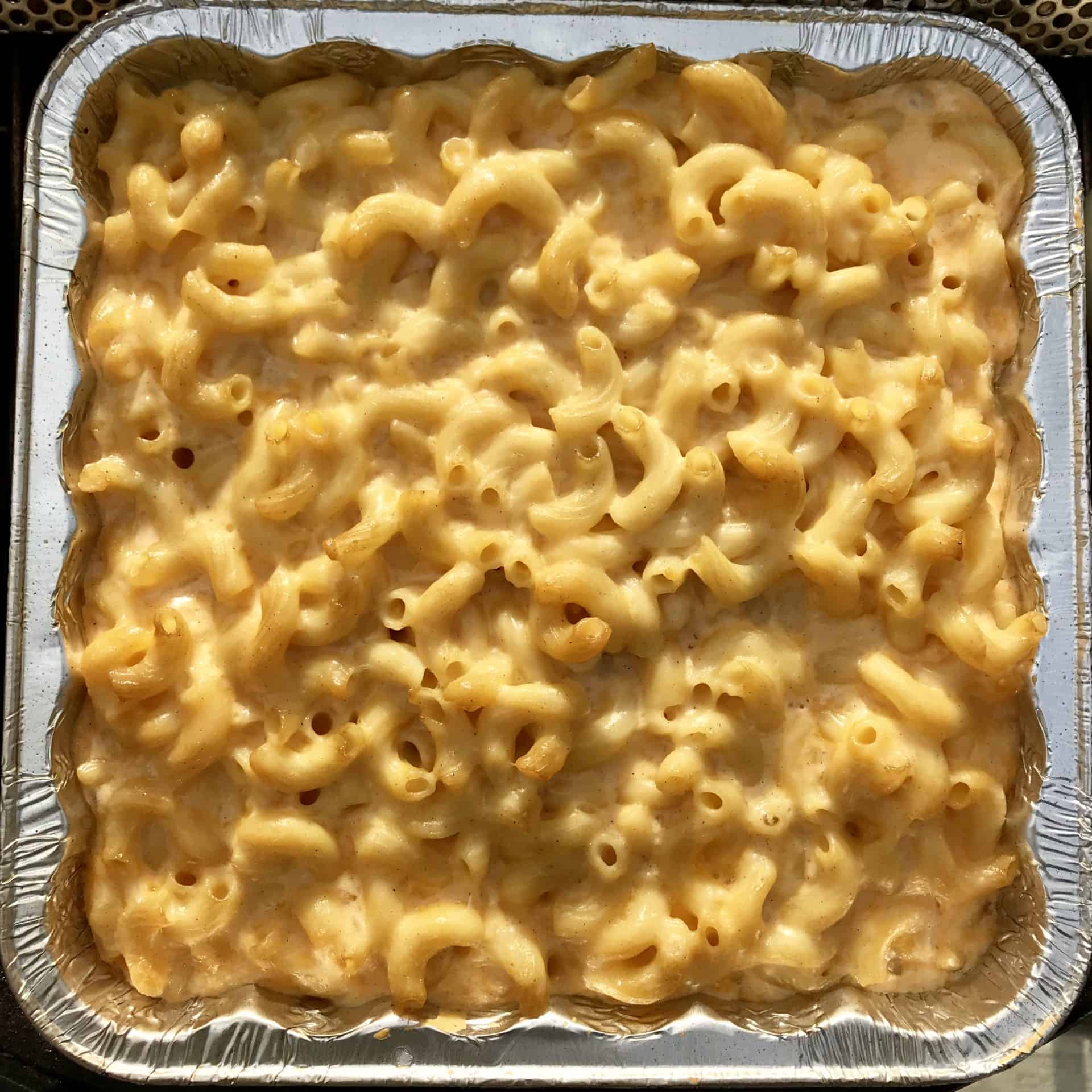 A close-up of baked macaroni and cheese in a square aluminum tray, showing golden, creamy pasta with a slightly browned cheesy top.