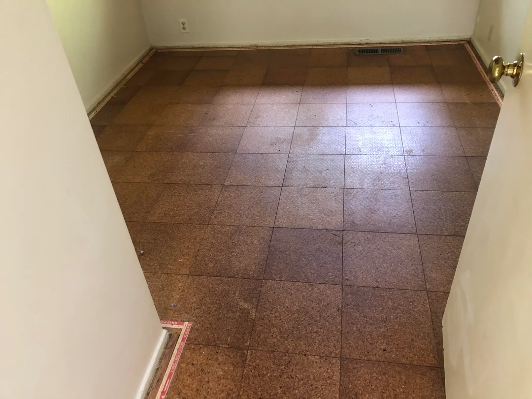 An empty room with brown square tiled flooring, cream-colored walls, and a white door partially open. There is a floor vent near the far wall and electrical outlets on the back wall.