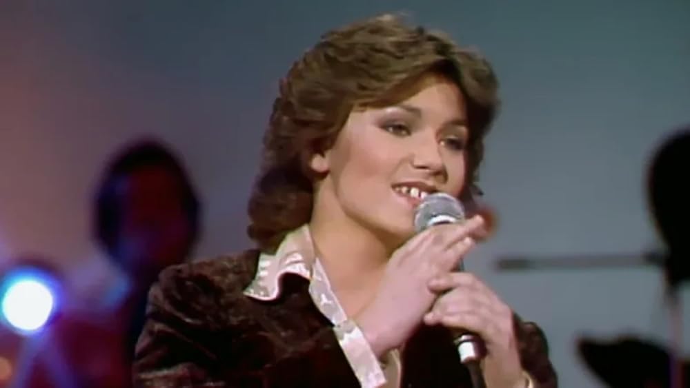 A person with short brown hair and wearing a dark jacket sings into a microphone on stage. The background is blurred with colorful lights and silhouettes of musicians.
