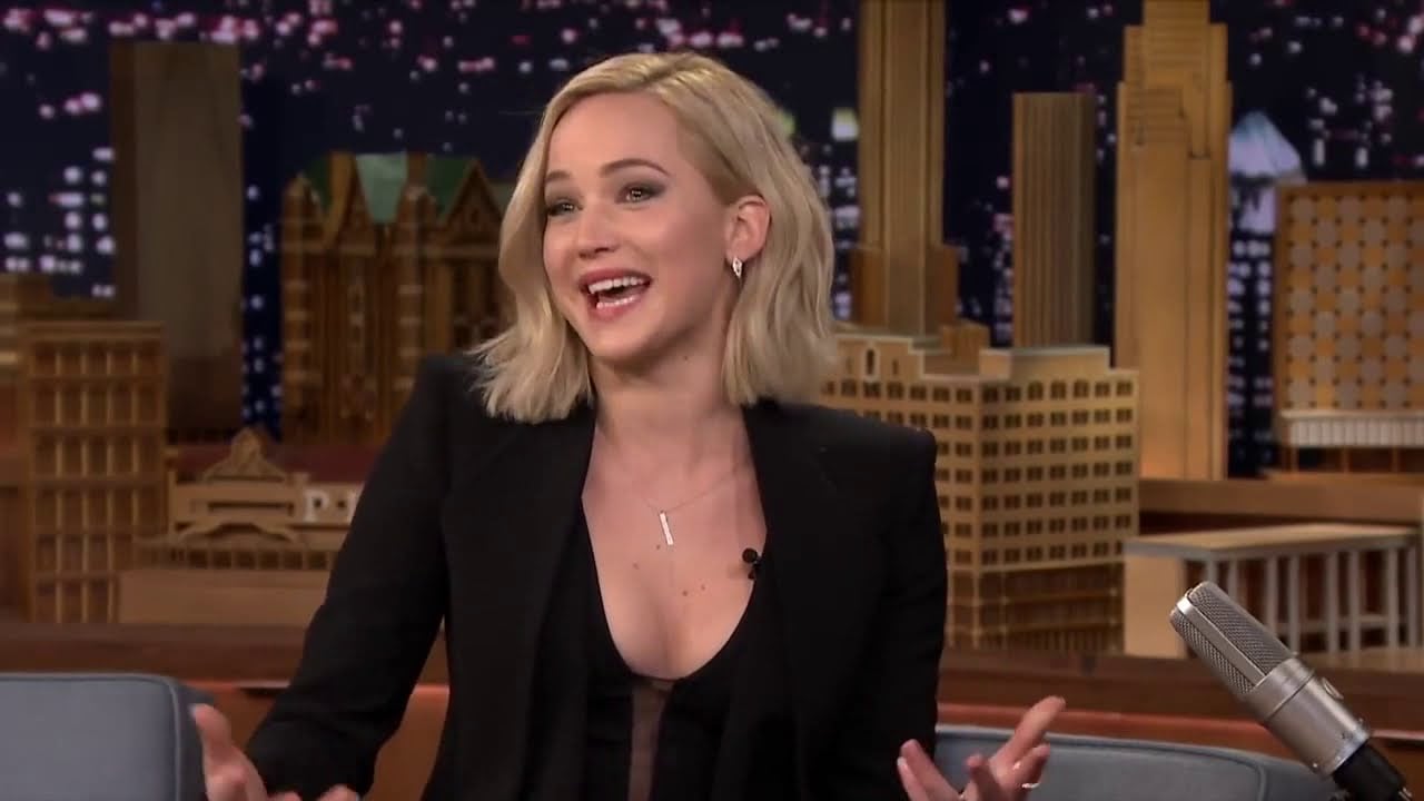 A woman with blonde, shoulder-length hair smiles and gestures with her hands while sitting on a talk show set, with a cityscape backdrop and a microphone visible in the foreground.