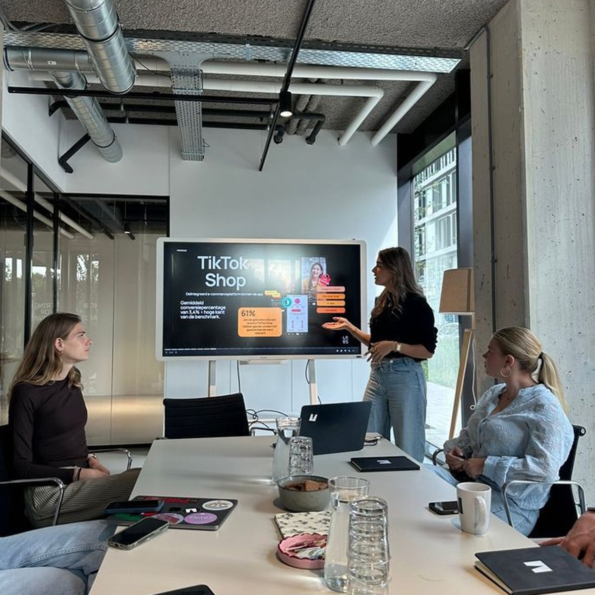 Four women are in a modern office meeting room. One is standing and presenting information about TikTok Shop on a large screen, while the others sit around a table with laptops, notebooks, and drinks.