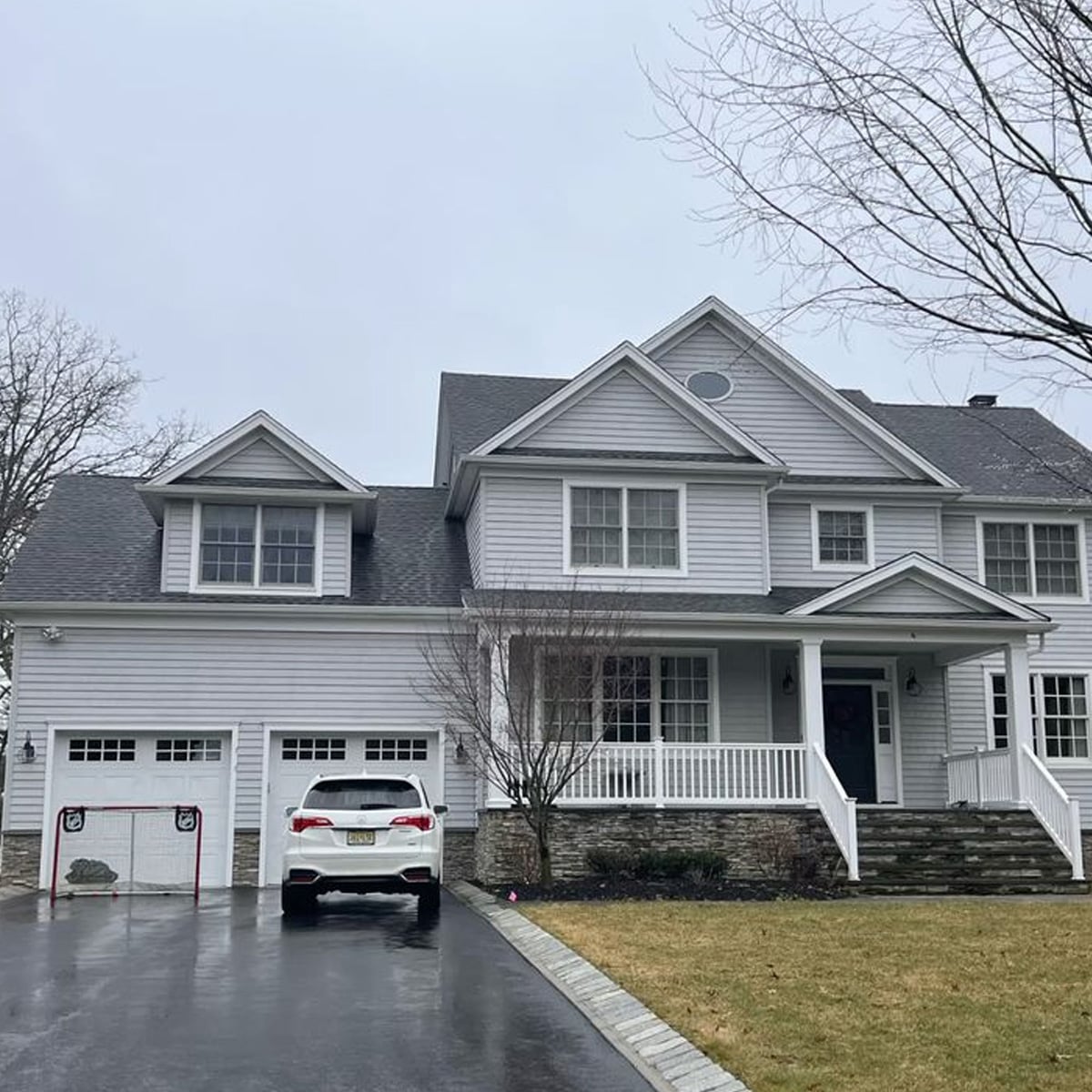 A large two-story gray house with white trim, an attached double garage, a white SUV parked in the driveway, and a front porch with stairs, on a cloudy day with bare trees and a patchy lawn.