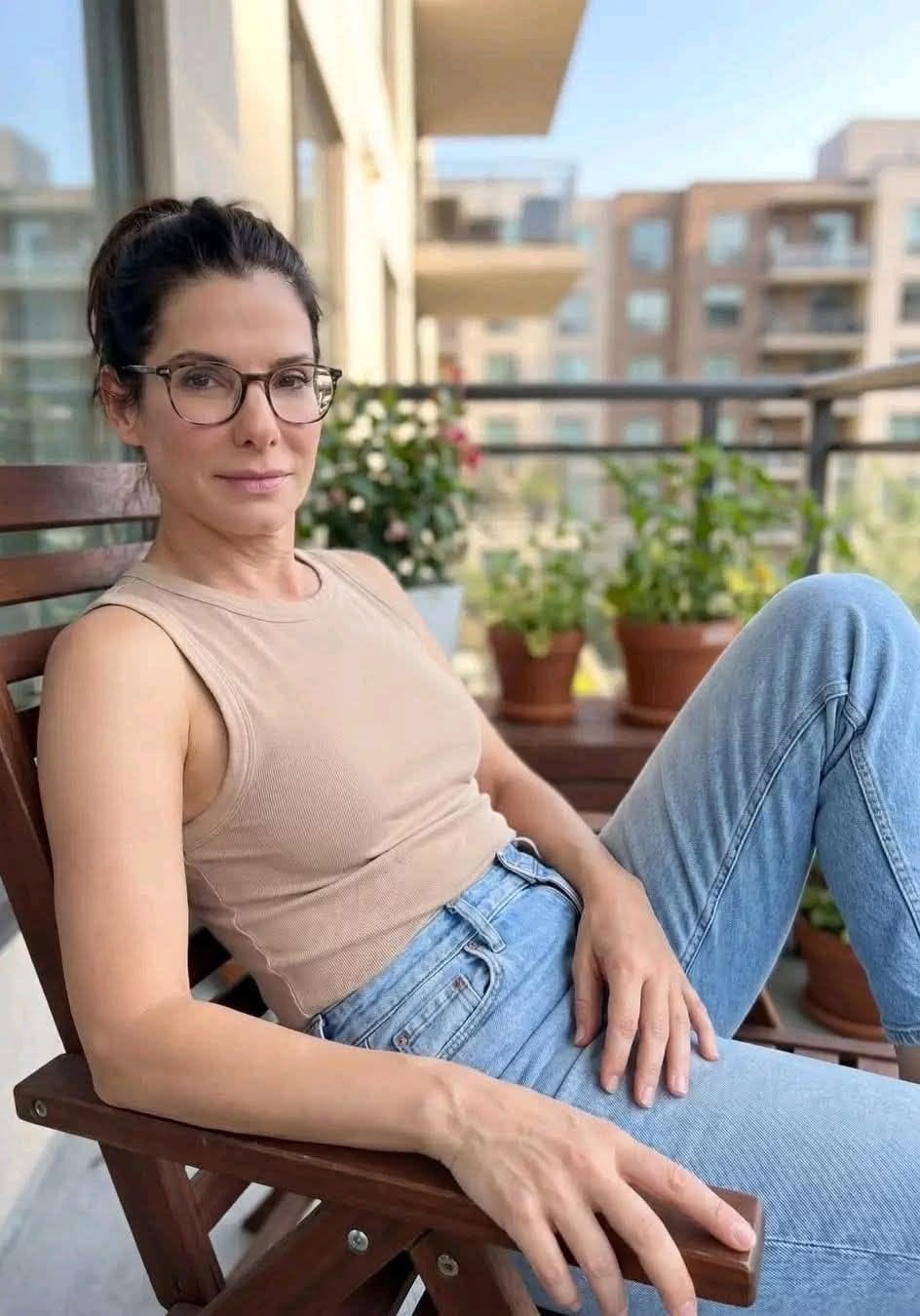 A woman wearing glasses, a beige sleeveless top, and blue jeans sits on a wooden chair on a balcony, with potted plants and modern buildings in the background.