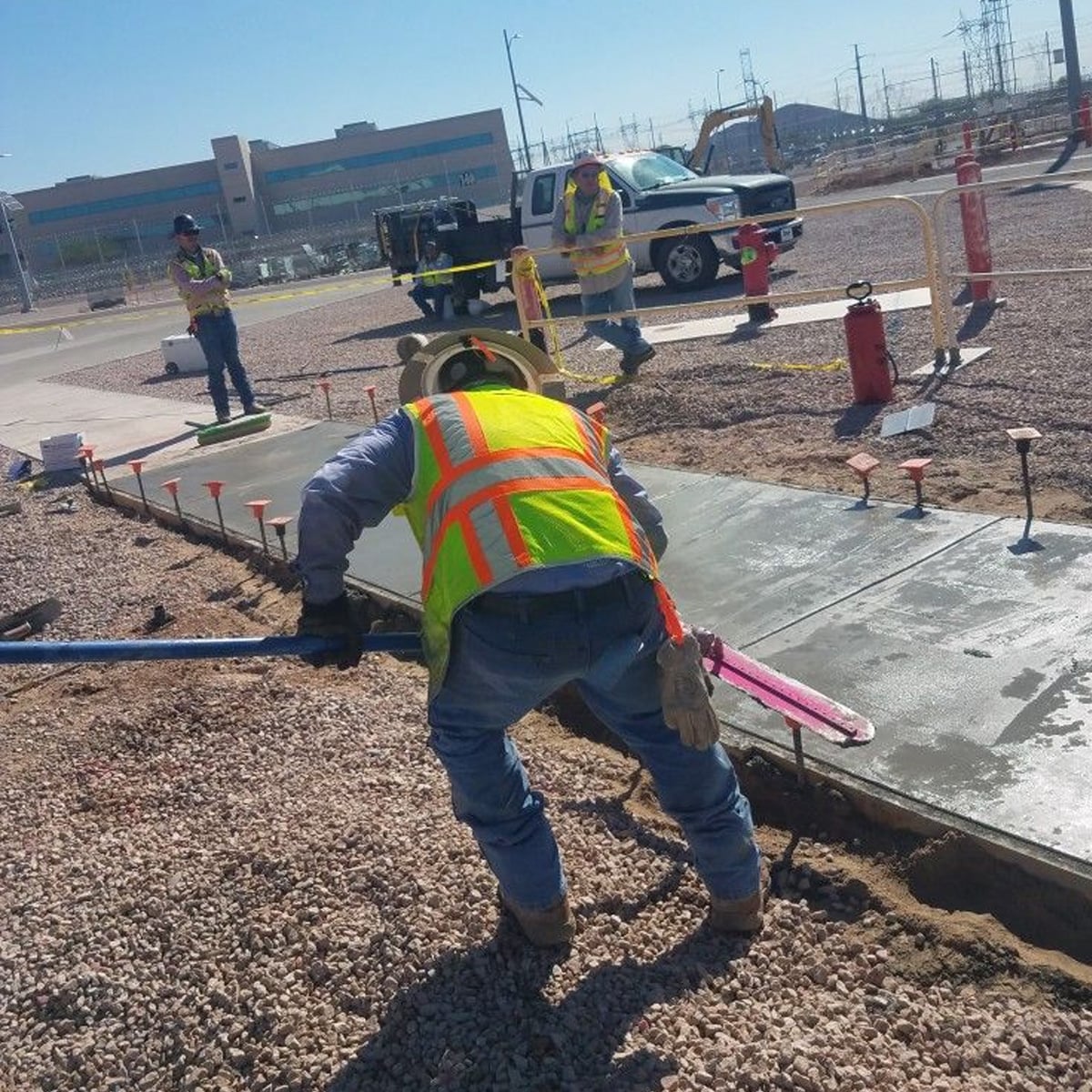 A construction worker in a neon safety vest smooths wet concrete on a new sidewalk while other workers and vehicles are visible in the background.