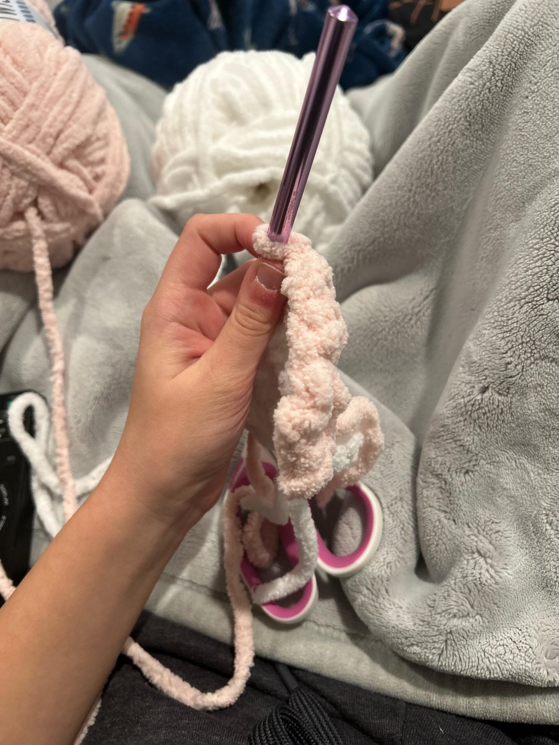 A hand holds a crochet hook with light pink yarn, working on a crochet project. Balls of soft pink and white yarn and a pair of pink-handled scissors rest on a gray blanket in the background.