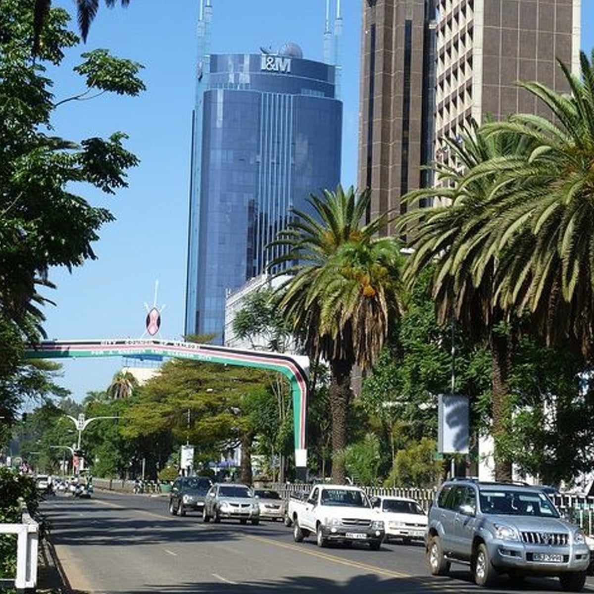 A city street lined with palm trees and tall buildings, with cars driving on the road. A blue glass skyscraper marked "I&M" stands prominently in the background, and a decorated arch spans the street.