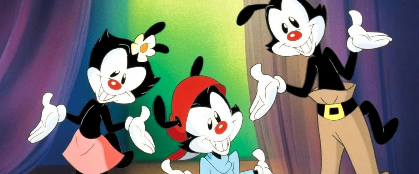 Three cartoon characters with black fur, white faces, and red noses pose happily in front of colorful curtains. One wears a skirt and flower, one a red cap, and one brown pants with a belt.