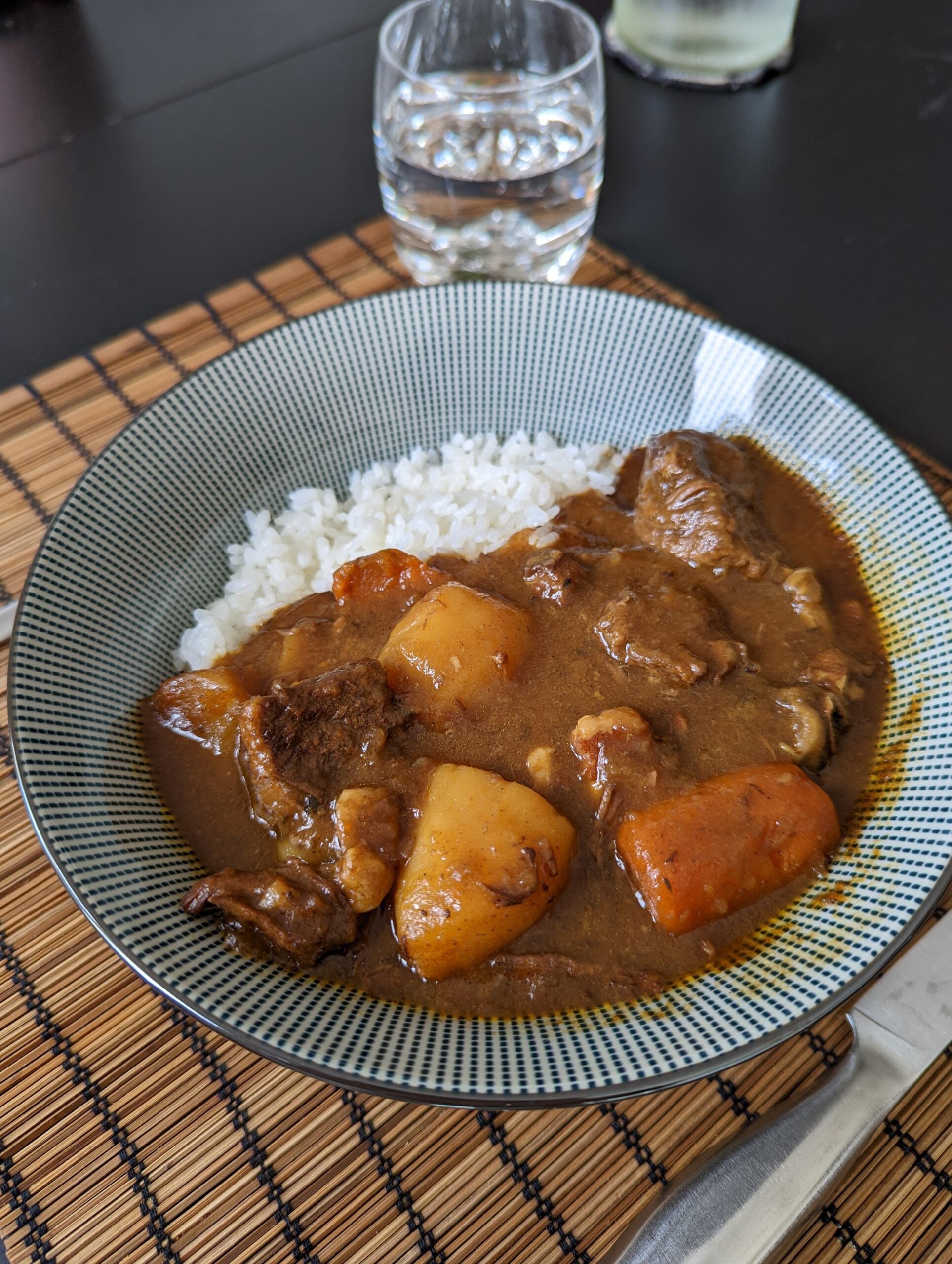 A bowl of Japanese curry with chunks of beef, potatoes, and carrots served alongside white rice, placed on a bamboo mat with a glass of water in the background.