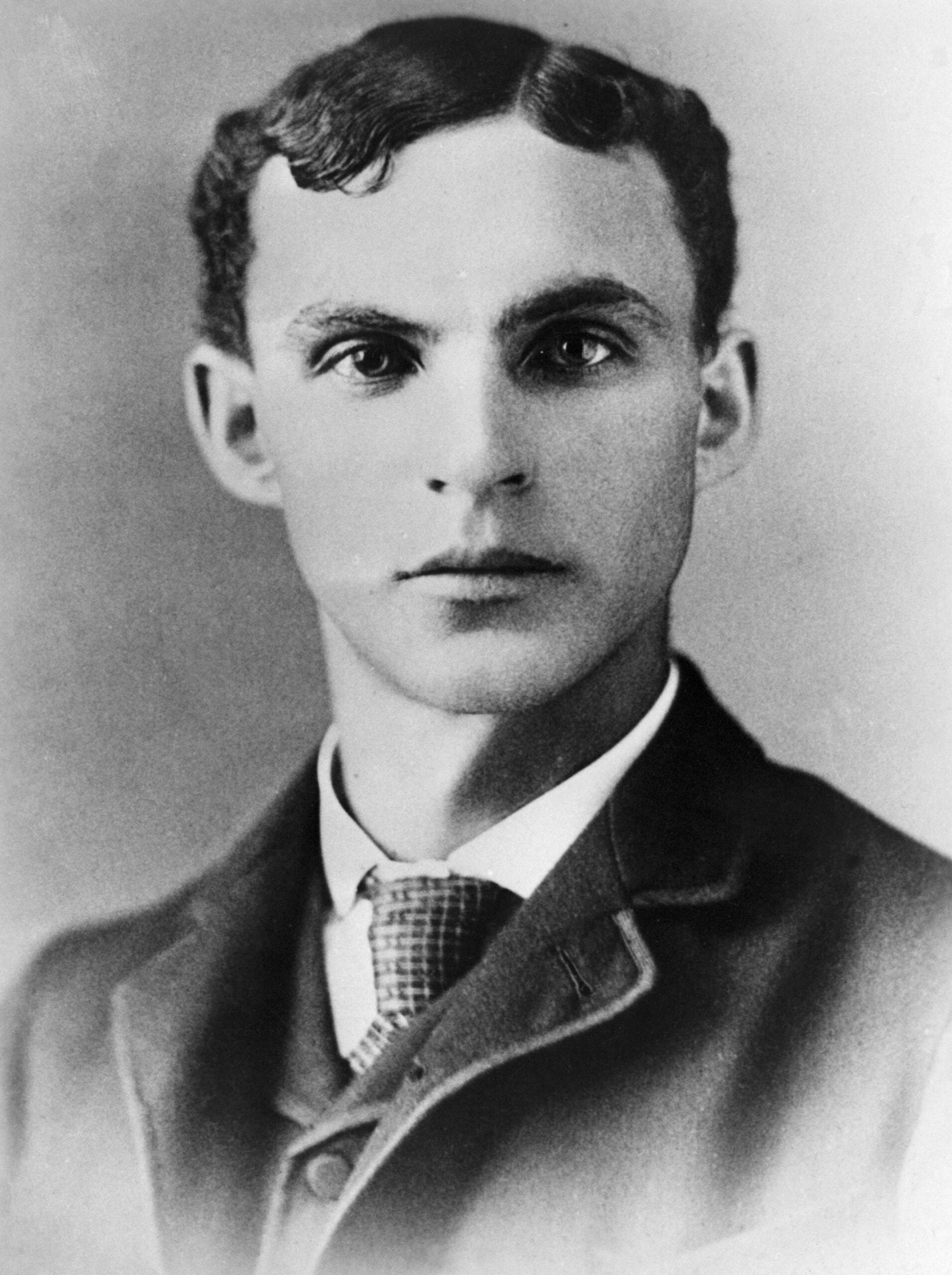 Black and white portrait of a young man with short, wavy hair, wearing a suit, vest, and patterned tie, looking directly at the camera with a neutral expression.
