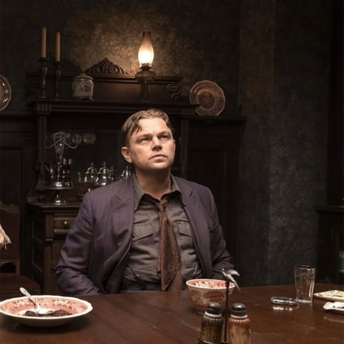 A man with slicked-back hair and a loosened tie sits at a dark wooden dining table with bowls of food, looking upward. The room has dim lighting and antique furniture in the background.