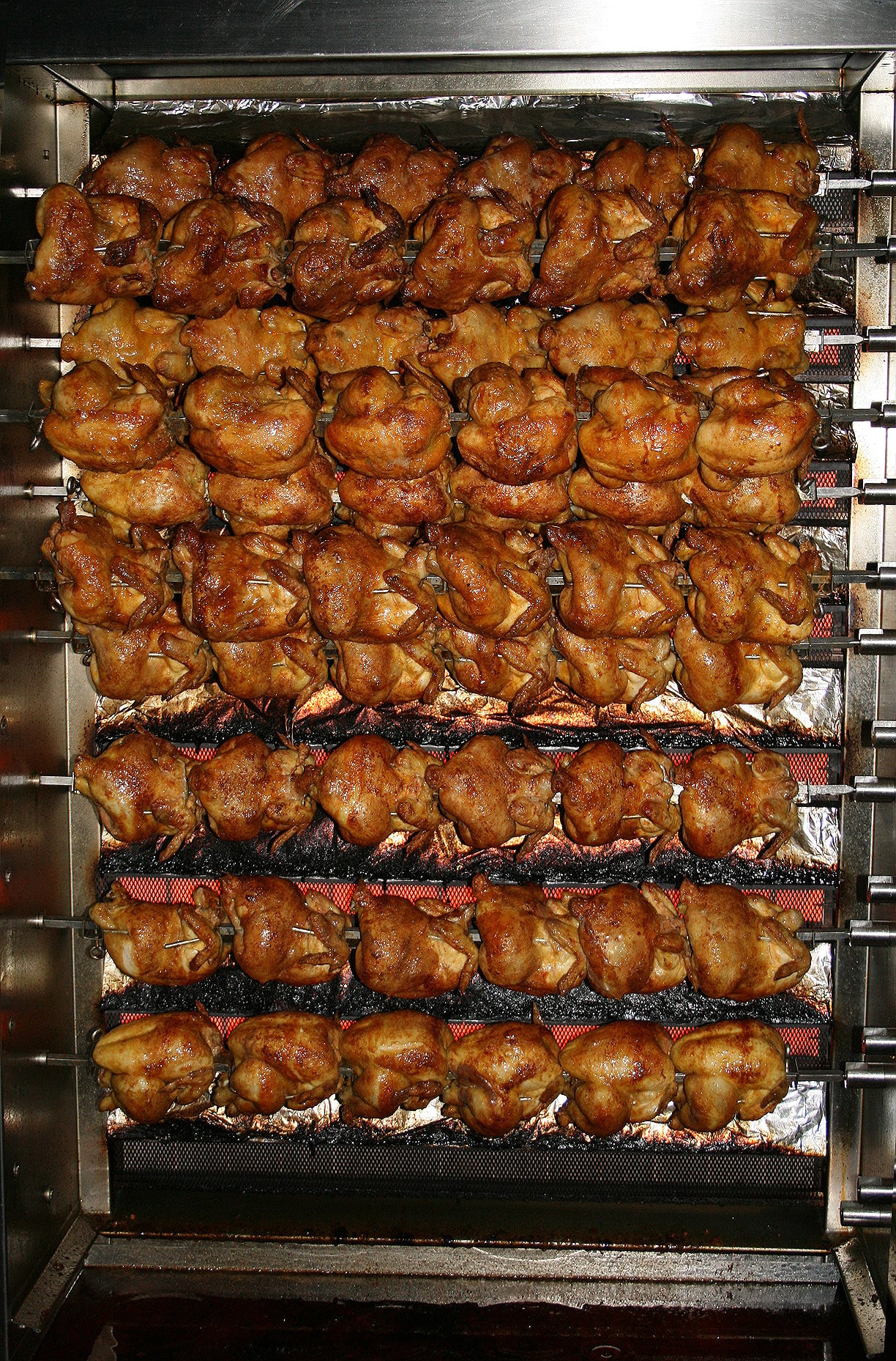 Rows of whole roasted chickens on a commercial rotisserie, golden brown and evenly cooked, with metal rods holding them in place above a drip tray.