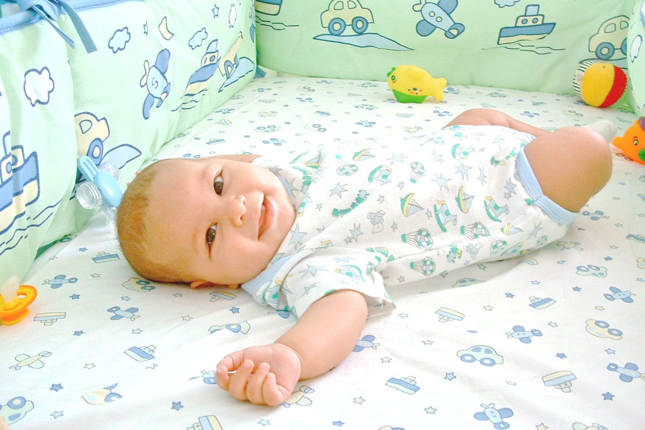 A baby wearing a printed onesie is lying on its back in a crib decorated with blue and green car and boat patterns, surrounded by soft toys and a pacifier.