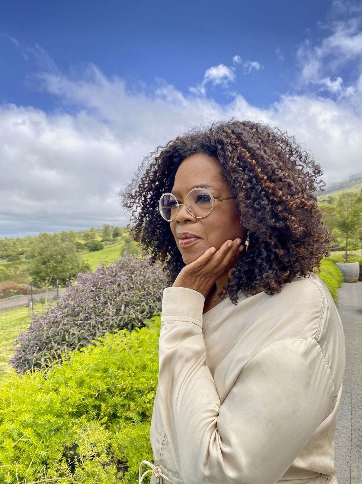 A woman with curly hair and round glasses stands outdoors, resting her hand on her chin and looking thoughtful. She wears a light-colored top, with green bushes and a bright, cloudy sky in the background.