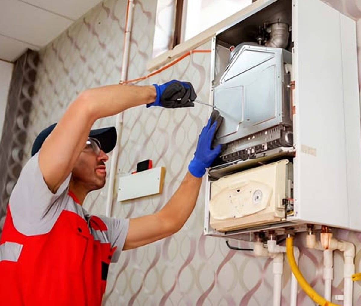 A technician wearing a red vest, gloves, and safety glasses repairs an open wall-mounted gas boiler, using a screwdriver to work on its internal components. Pipes and wires are visible below the boiler unit.