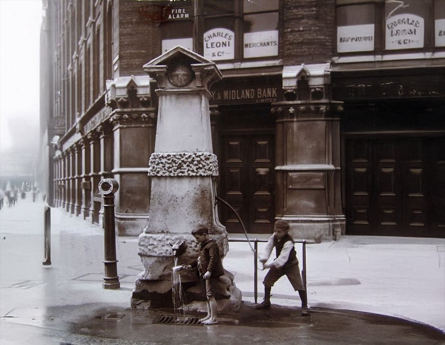 Two boys use a public water fountain on a city street corner near a large stone building; one drinks while the other pumps water. Signs for businesses and a bank are visible in the background. The scene appears historical.